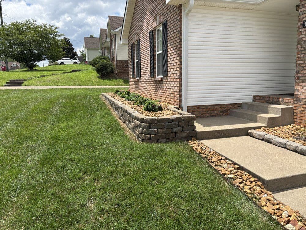 Raised flower bed with stacked retaining wall blocks and river rock beside a front walk